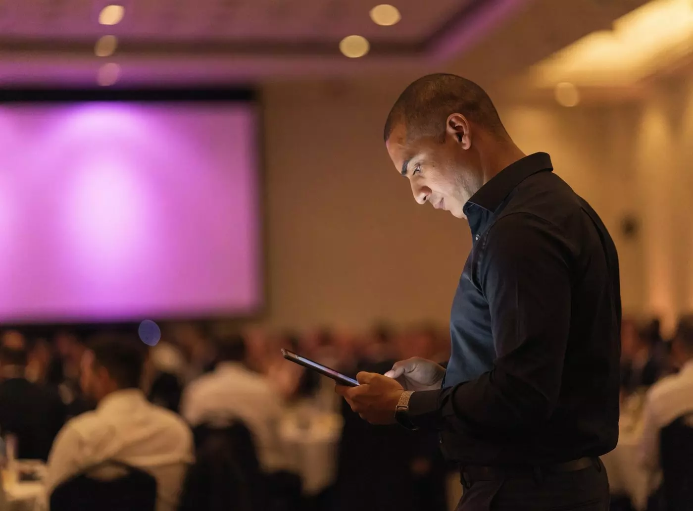 Master of ceremonies running the order of service from a tablet at the side of the room, large screen lit in the background