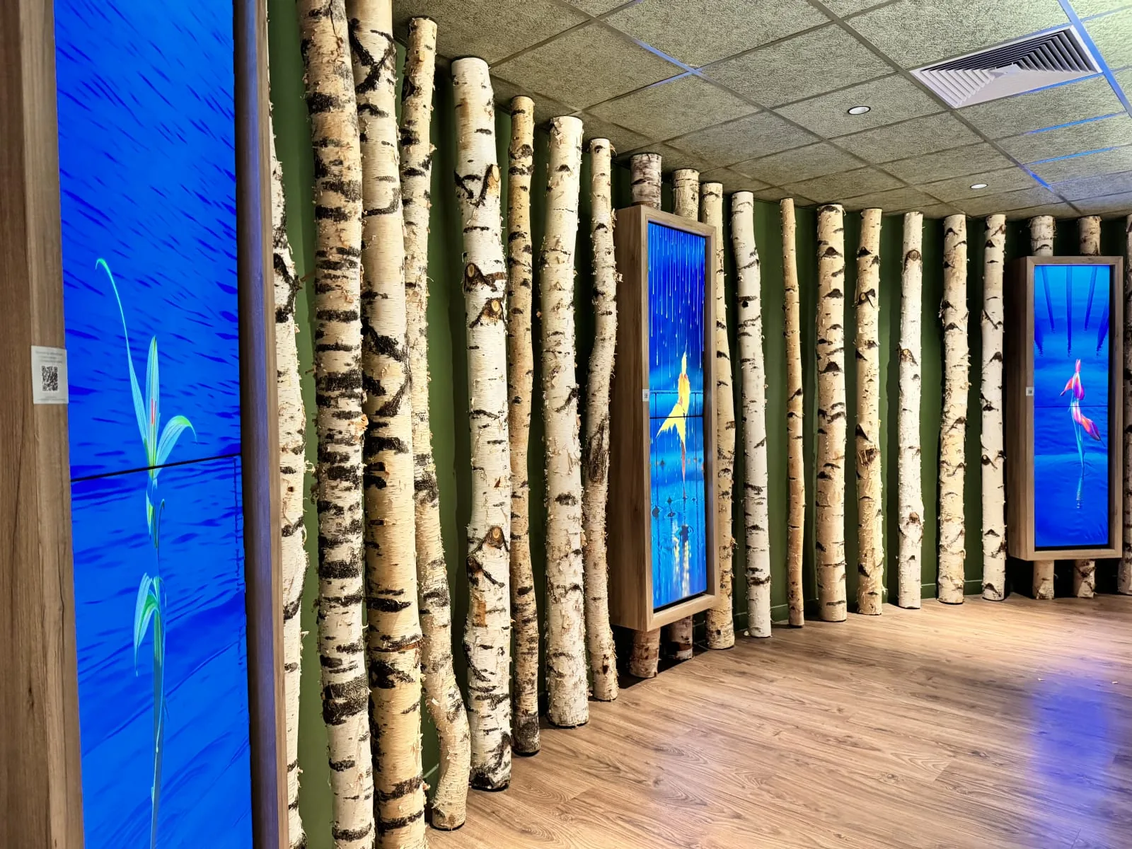 Couloir d&rsquo;h&ocirc;tel bord&eacute; de troncs de bouleau, o&ugrave; des &eacute;crans verticaux diffusent des &oelig;uvres bleues