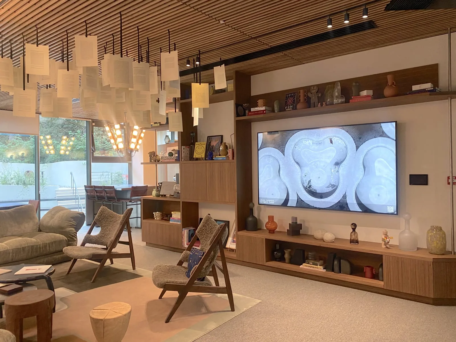 Grand &eacute;cran diffusant une &oelig;uvre sculpturale blanche dans un espace de d&eacute;tente meubl&eacute; de fauteuils en bois