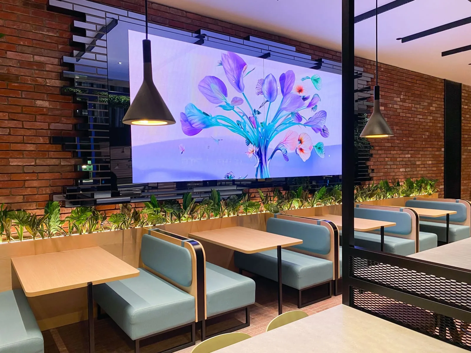 Mur LED diffusant une &oelig;uvre florale au-dessus des banquettes d&rsquo;un restaurant d&rsquo;entreprise, entre brique apparente et plantes