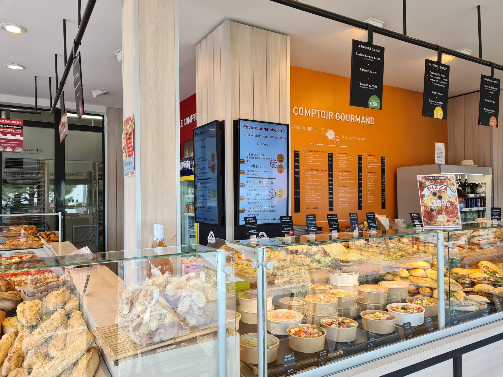 Taulell d'una fleca Atelier Banette: dues pantalles presenten els entrepans disponibles, sota les ofertes del migdia mostrades sobre la vitrina refrigerada
