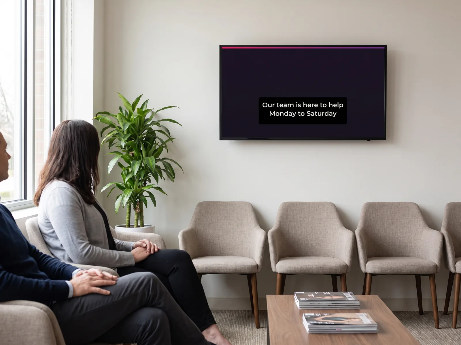 Wall-mounted screen in a waiting area playing a subtitled video, two people seated watching it