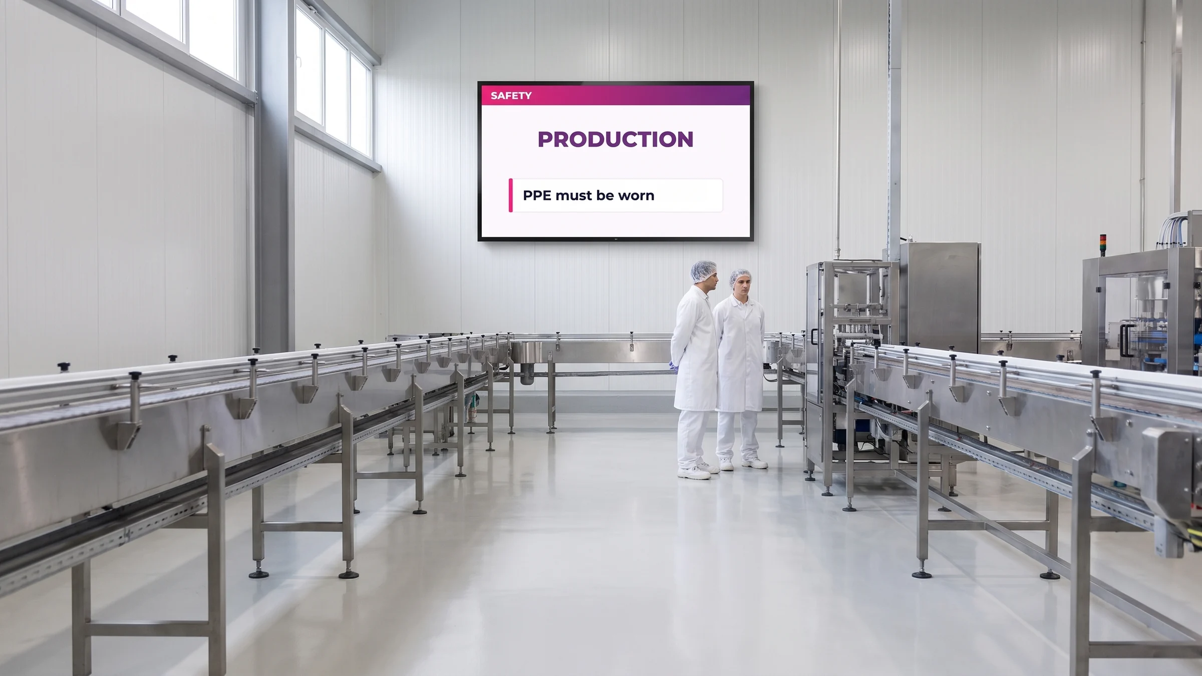 Wall-mounted screen showing a safety notice at the end of a stainless-steel packaging line, with two operators in white coats in the foreground