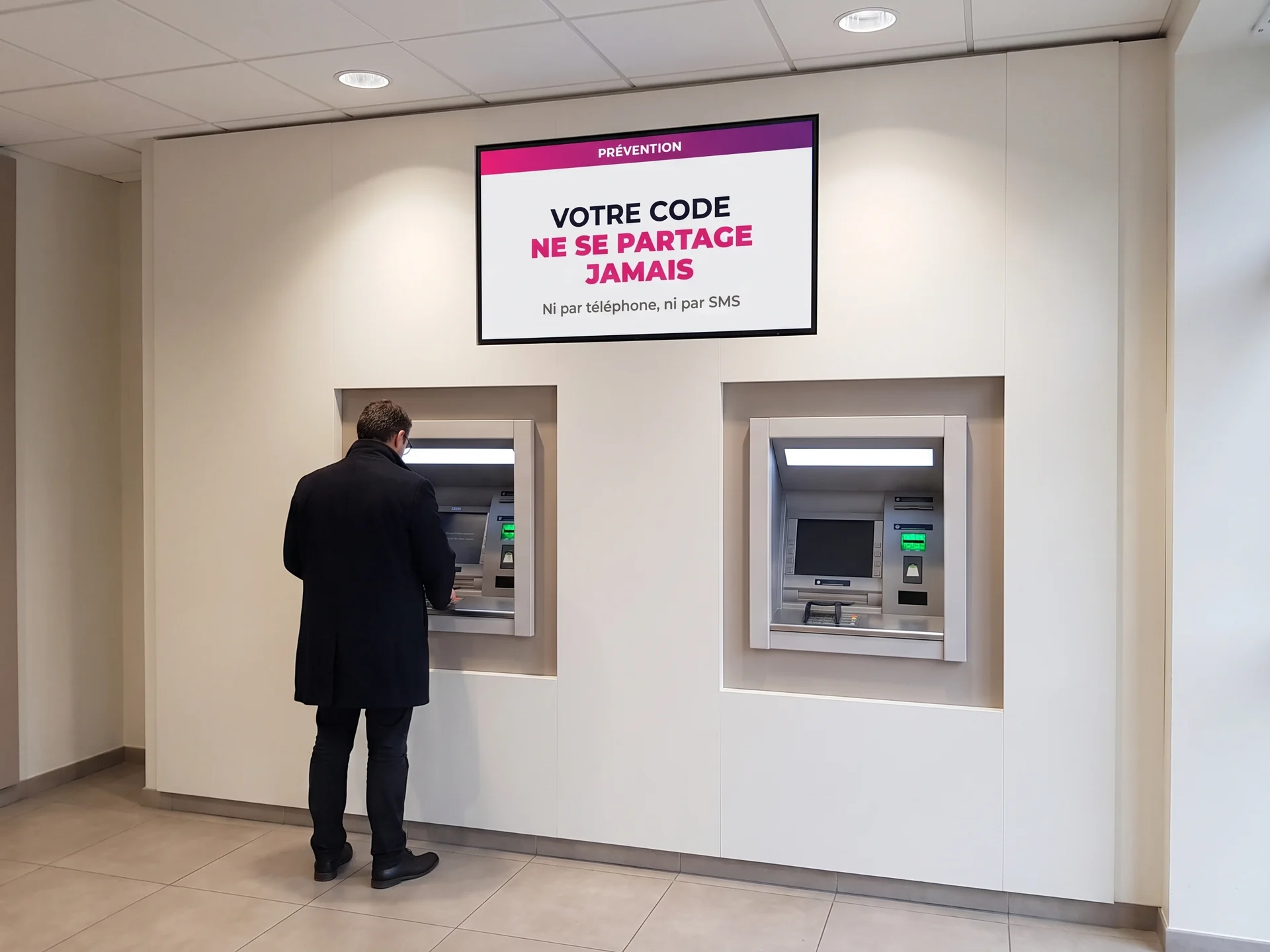 Espace libre-service bancaire avec deux automates et un &eacute;cran de pr&eacute;vention rappelant de ne jamais communiquer son code
