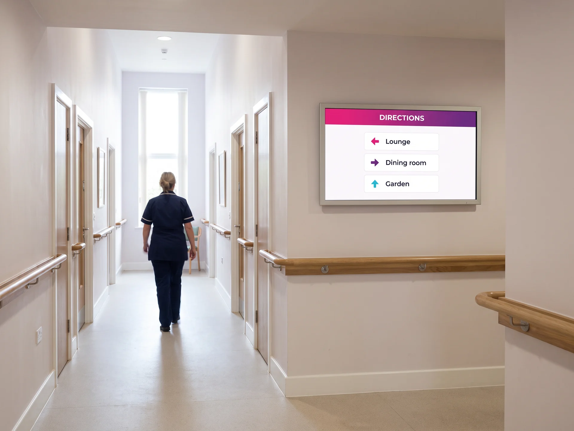 Wayfinding screen at a care home corridor junction, pointing to the lounge, the dining room and the garden