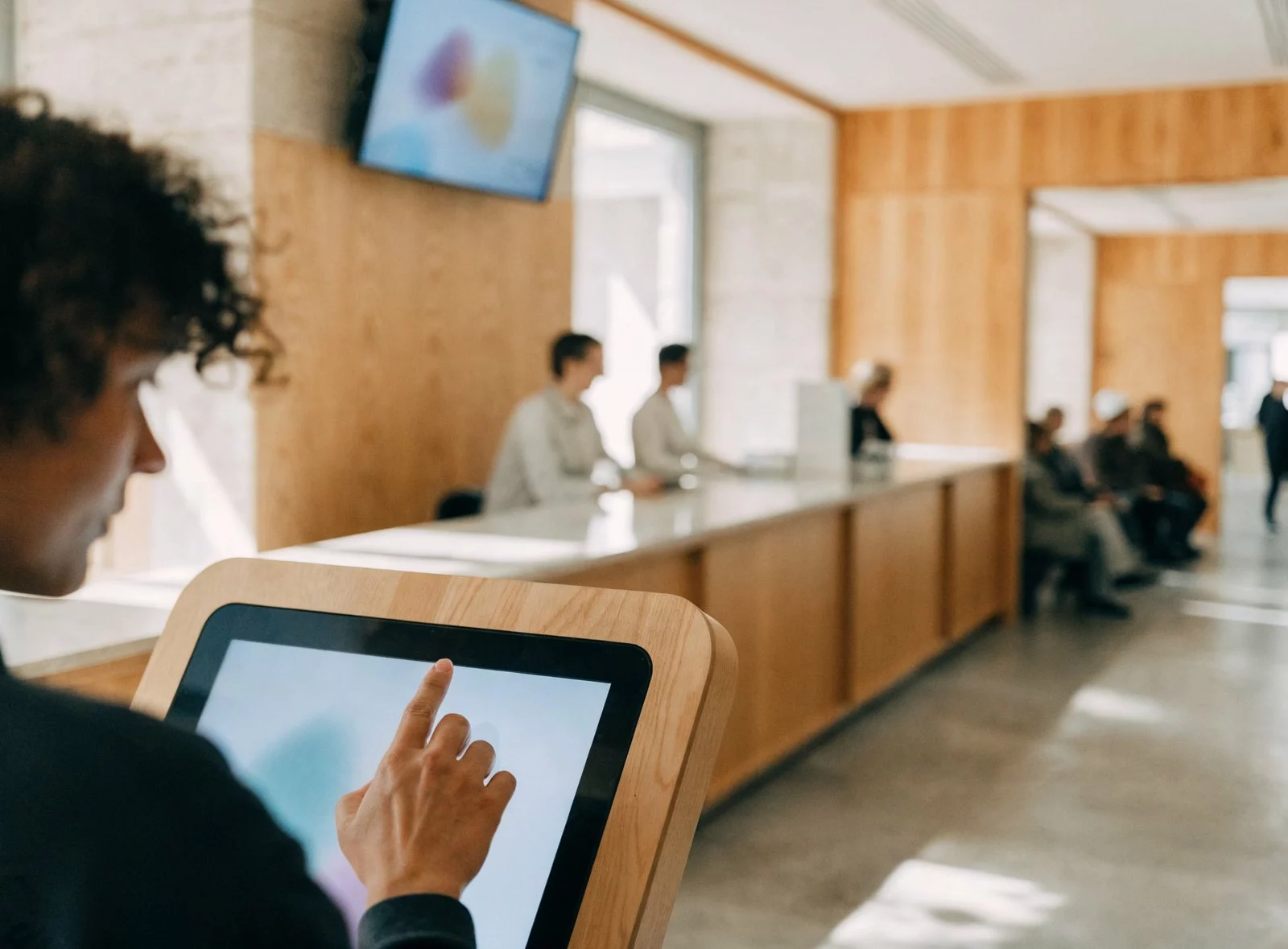 Resident using a touch screen kiosk in a town hall reception area, with an information screen above the counters