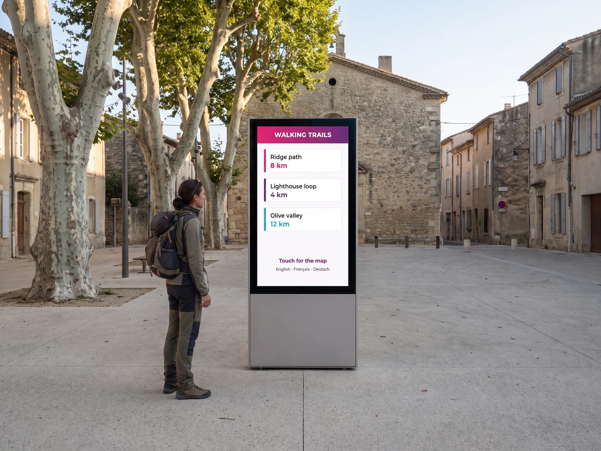Outdoor touchscreen information kiosk on a village square, listing walking trails and their distances in several languages