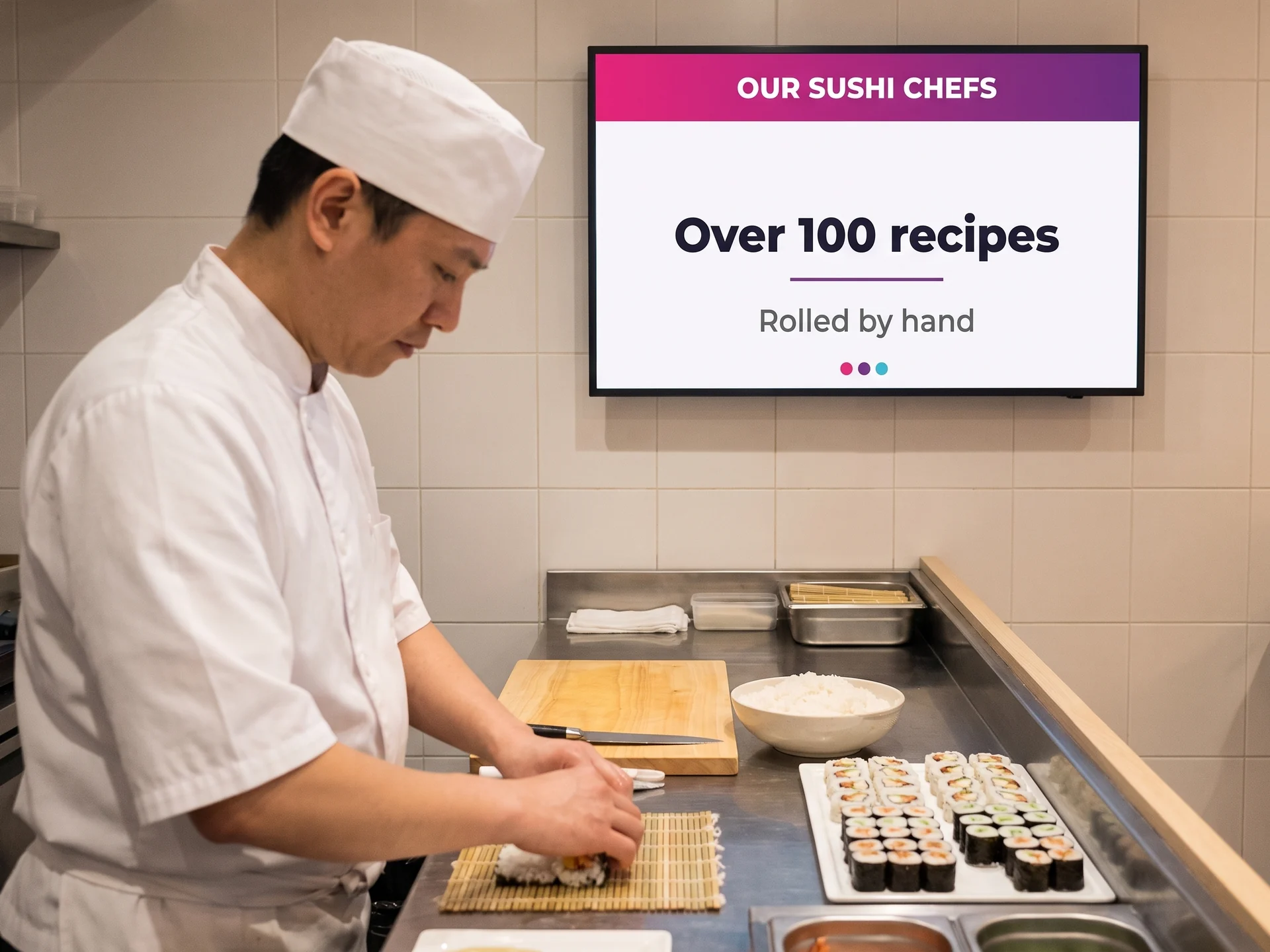 Chef rolling maki by hand behind a counter, wall screen above