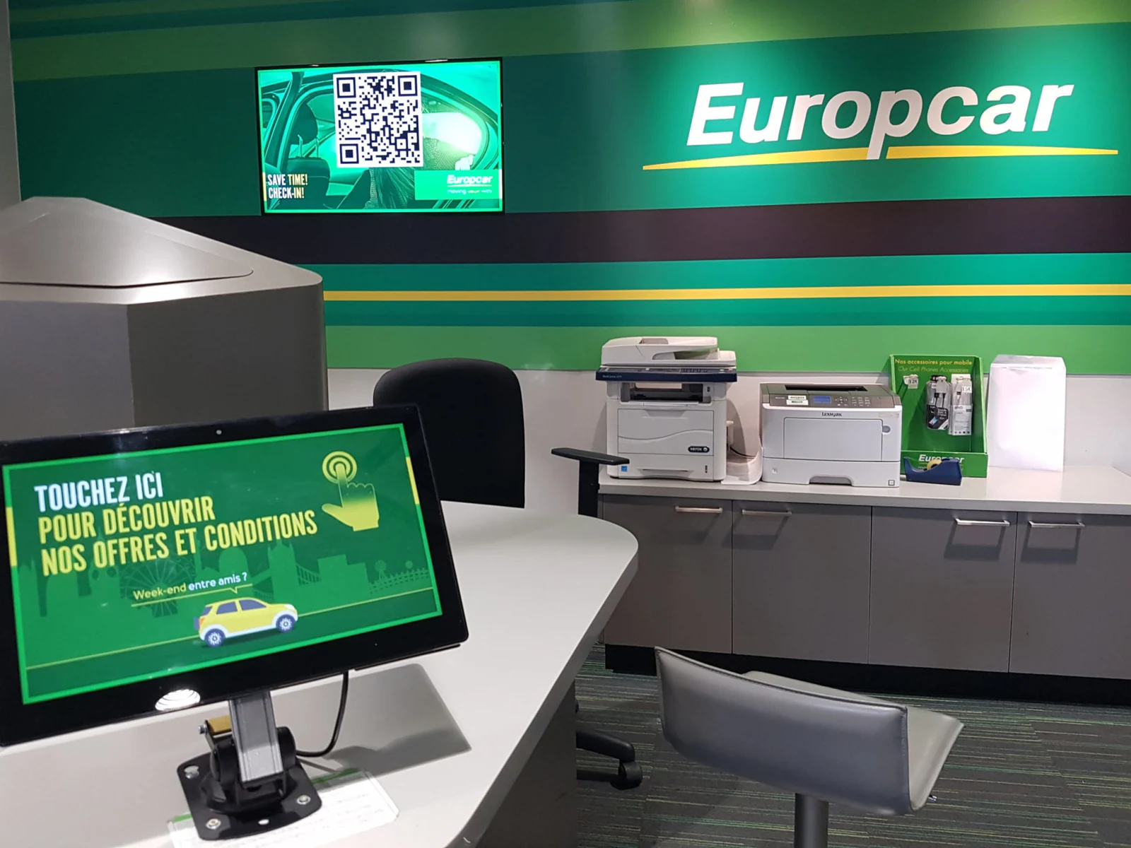 Comptoir d'une agence Europcar : écran mural diffusant une invitation au pré-enregistrement et tablette tactile posée sur le comptoir