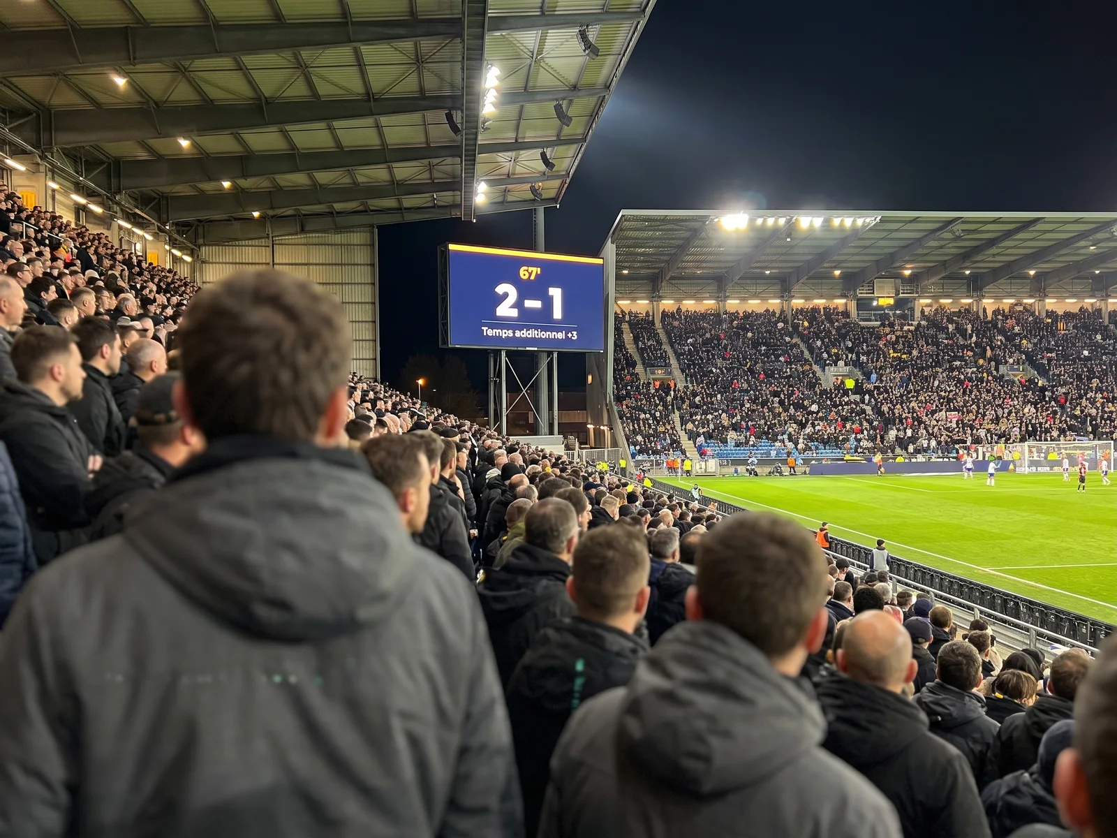 Spectateurs en tribune devant un écran géant affichant le score et le temps additionnel