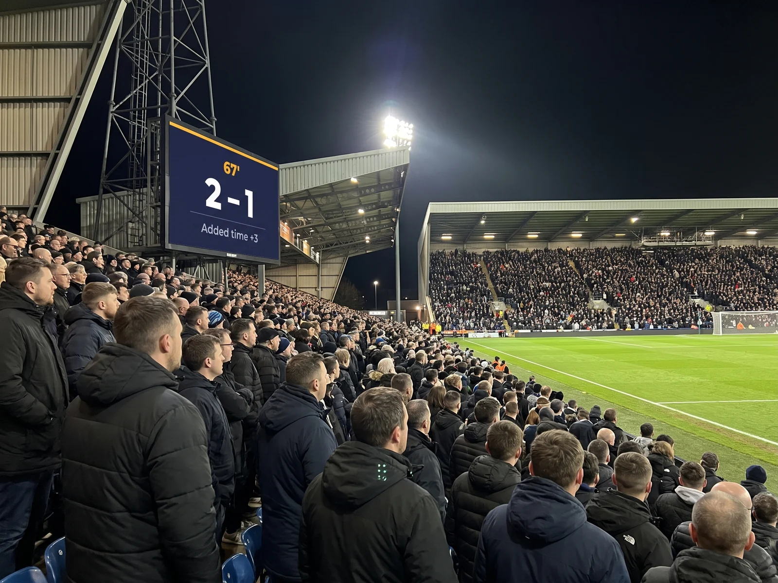 Supporters in a packed stand watching a giant screen showing the score and added time
