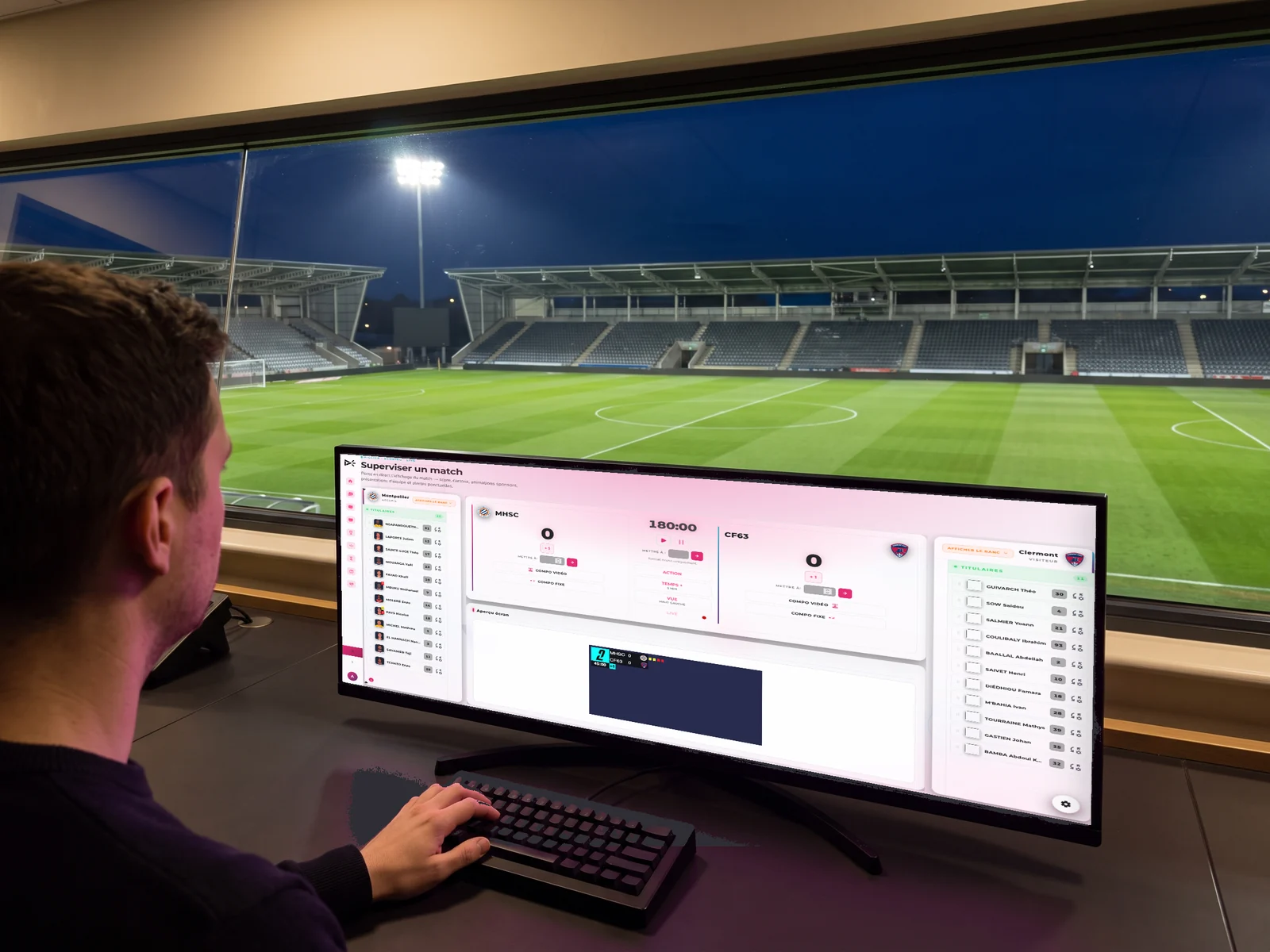 Poste de r&eacute;gie face &agrave; une baie panoramique sur la pelouse &eacute;clair&eacute;e, le moniteur affichant l'interface SCORING d'EMITY avec le score, le chronom&egrave;tre et les compositions d'&eacute;quipes