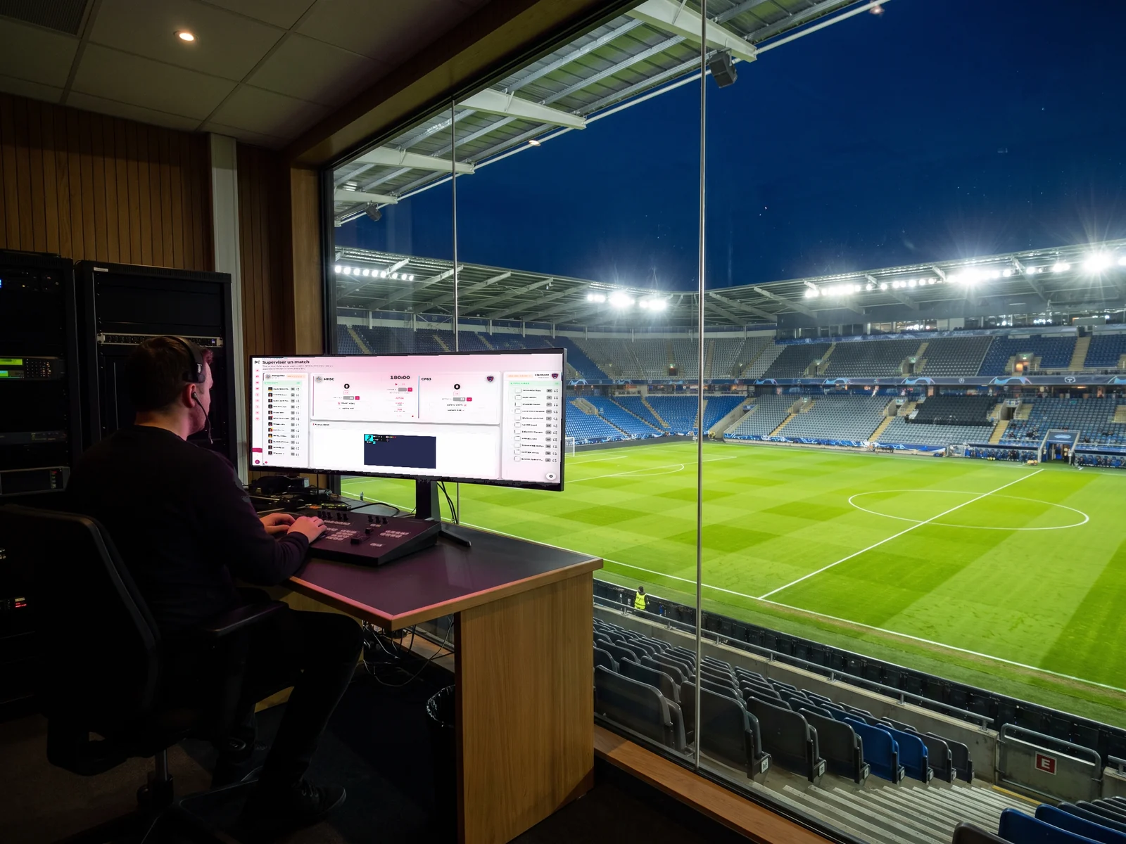 Opérateur en régie de stade devant une baie vitrée donnant sur la pelouse éclairée, son moniteur affichant l'interface SCORING d'EMITY