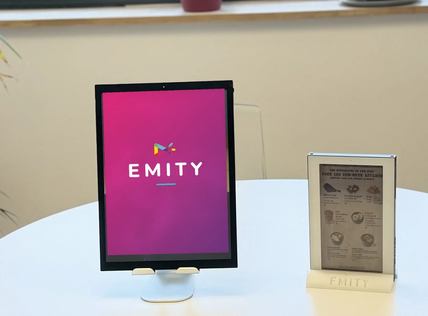 Écran e-paper de 13,3 pouces posé sur une table dans les bureaux EMITY, affichant un visuel aux couleurs de la marque à côté d'un chevalet papier