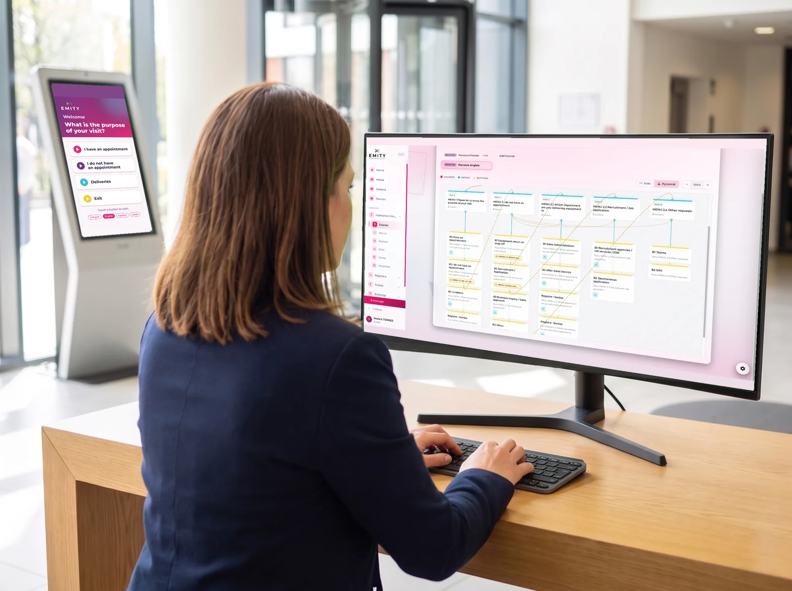 Reception manager building an interactive journey in E-MANAGER on a large monitor, in front of a touchscreen kiosk