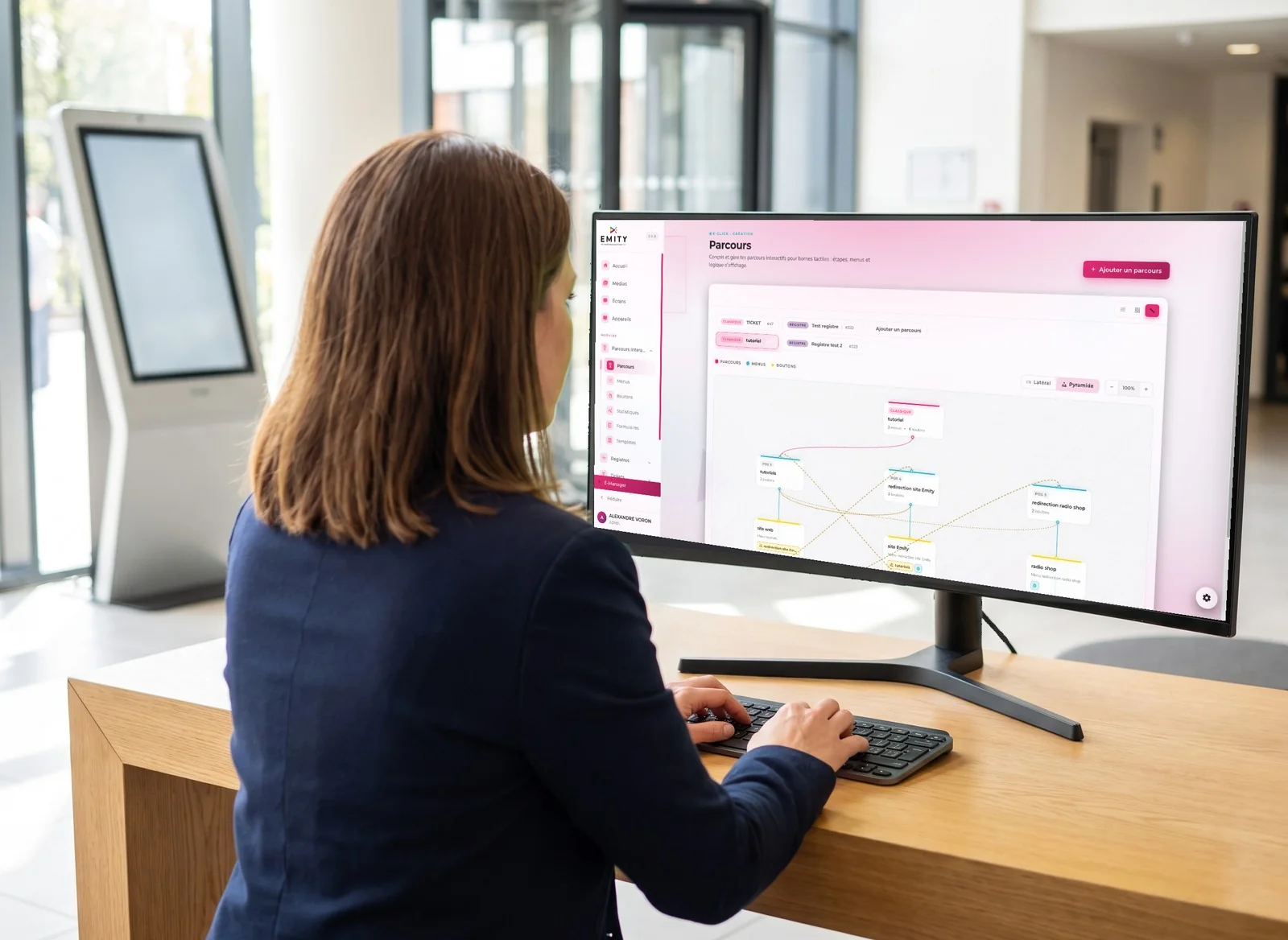 Responsable d'accueil qui compose un parcours interactif dans E-MANAGER sur un grand moniteur, devant une borne tactile