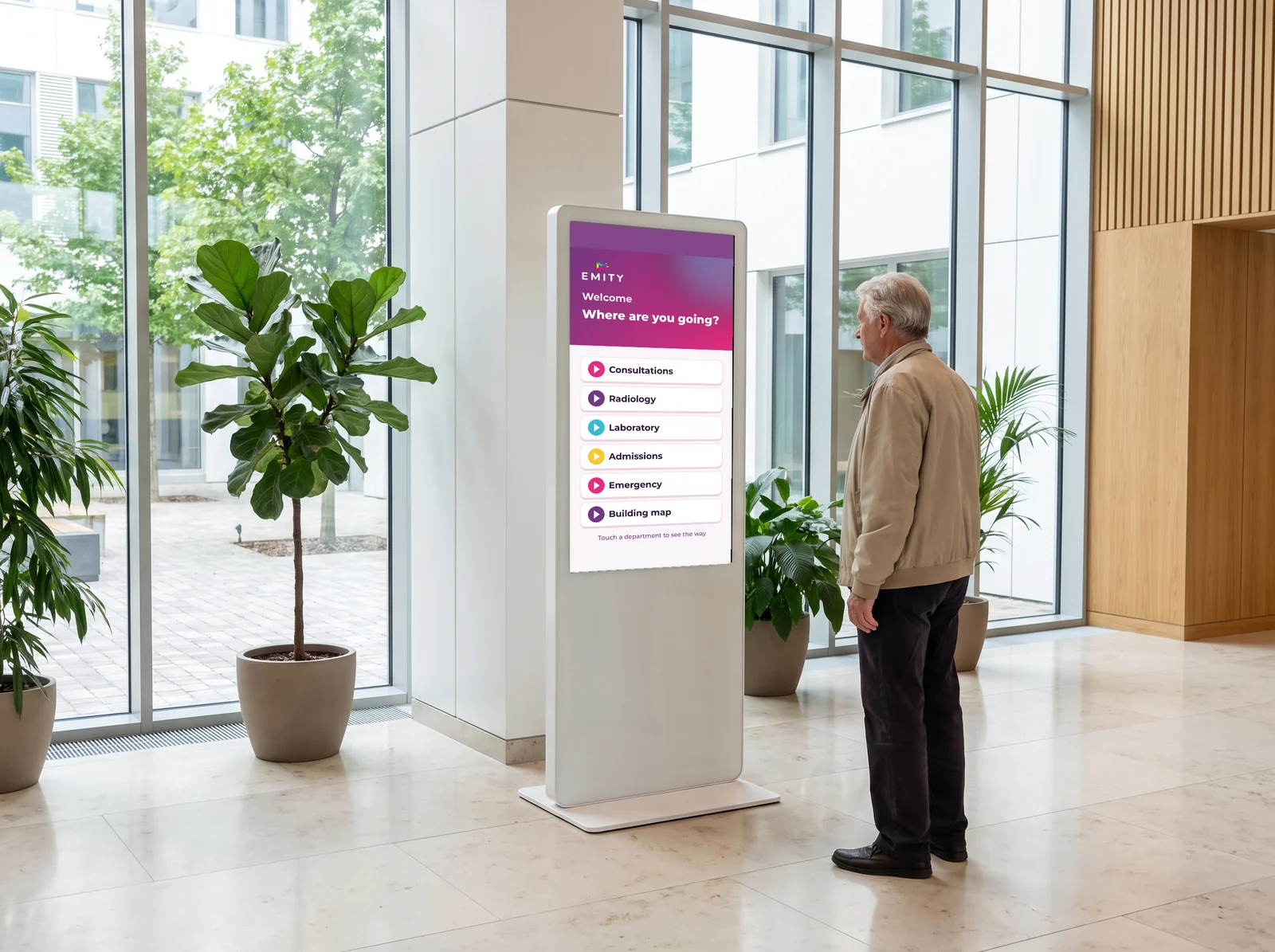 Patient at an E-CLICK touchscreen kiosk showing “Where are you going?” in a healthcare facility lobby: consultations, radiology, laboratory, admissions, emergency, building map