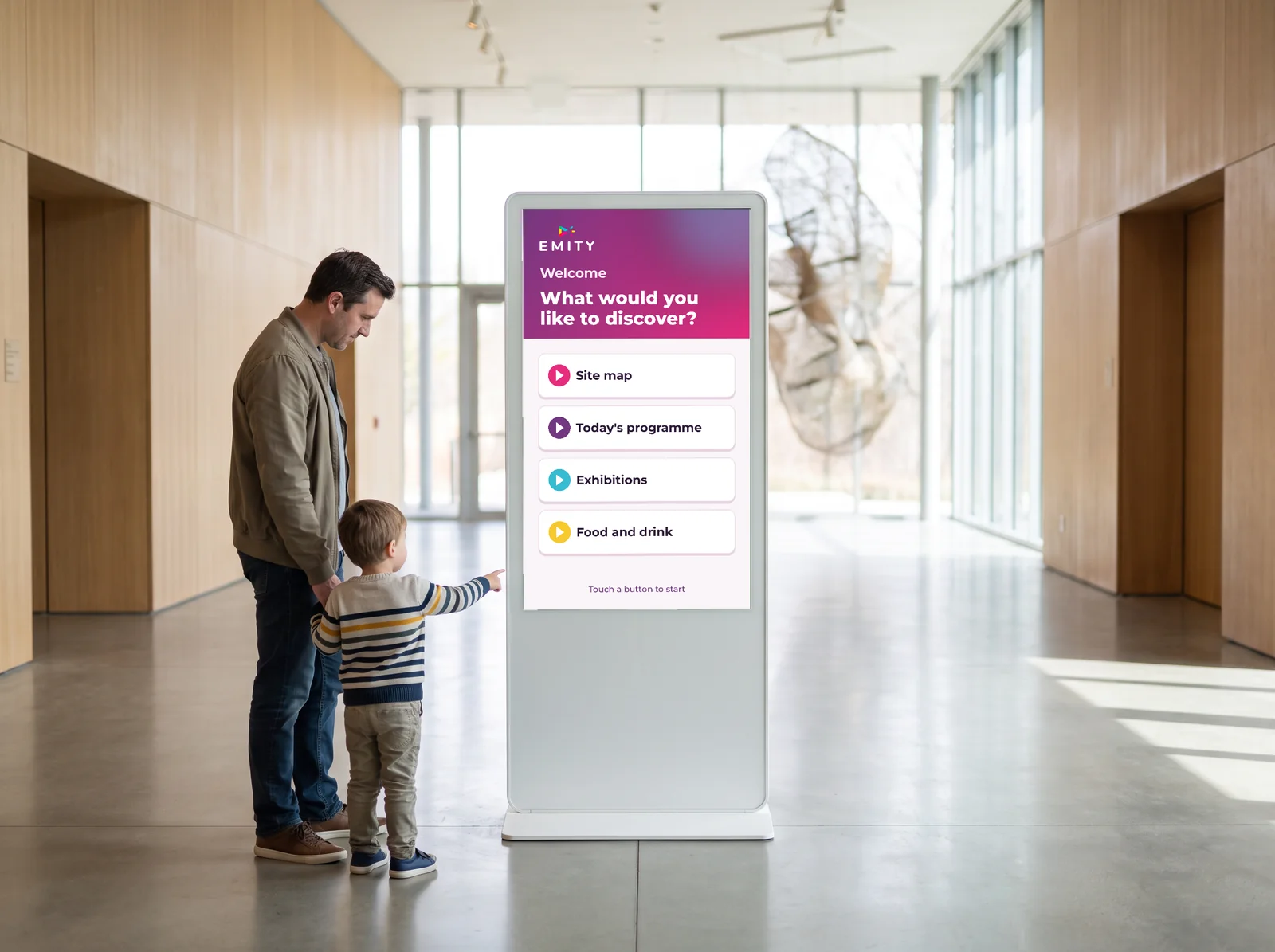 Father and child at an E-CLICK touchscreen kiosk showing “What would you like to discover?” in a museum lobby: site map, today's programme, exhibitions, food and drink