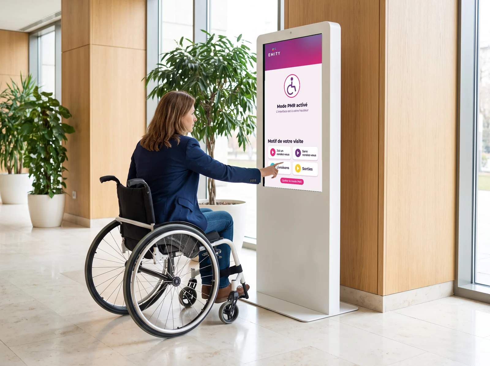 Femme en fauteuil roulant qui touche le bas d’une borne E-CLICK en mode PMR : l’interface « Motif de votre visite » est descendue à sa hauteur