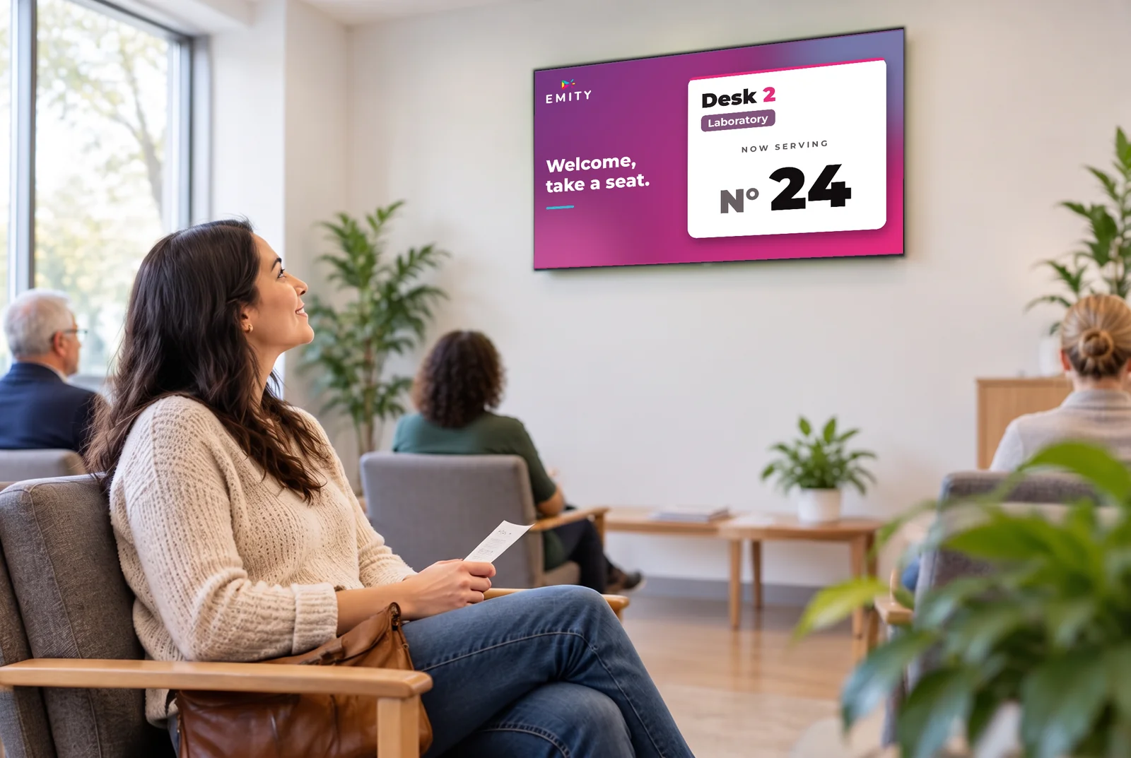 Seated visitor holding a ticket in a waiting room, in front of a call screen showing number 24 at desk 2
