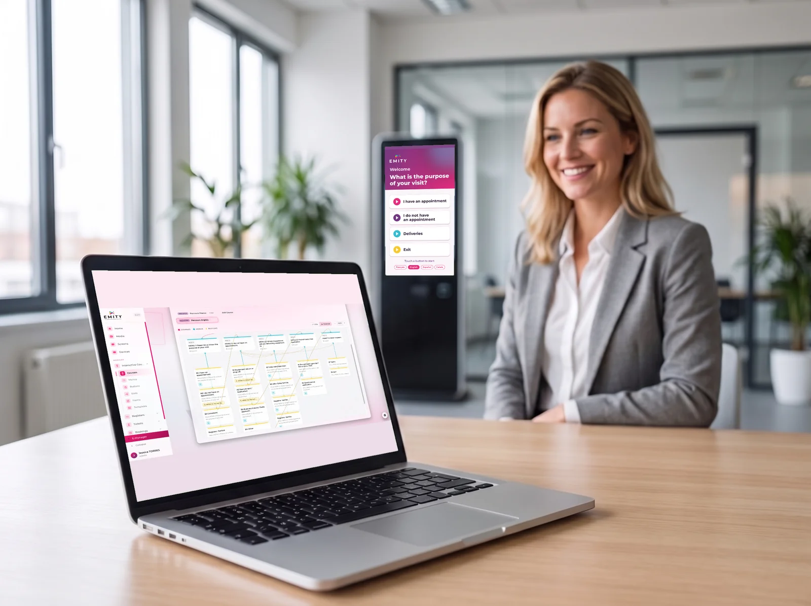 Smiling manager at a laptop open on the E-CLICK journey in E-MANAGER, a touchscreen kiosk in the background