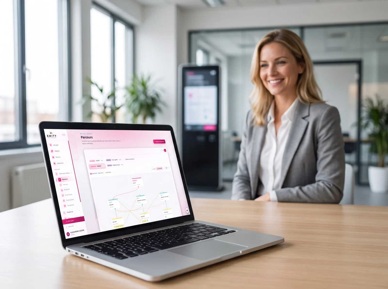 Responsable souriante devant un portable ouvert sur le parcours E-CLICK, une borne tactile en arrière-plan