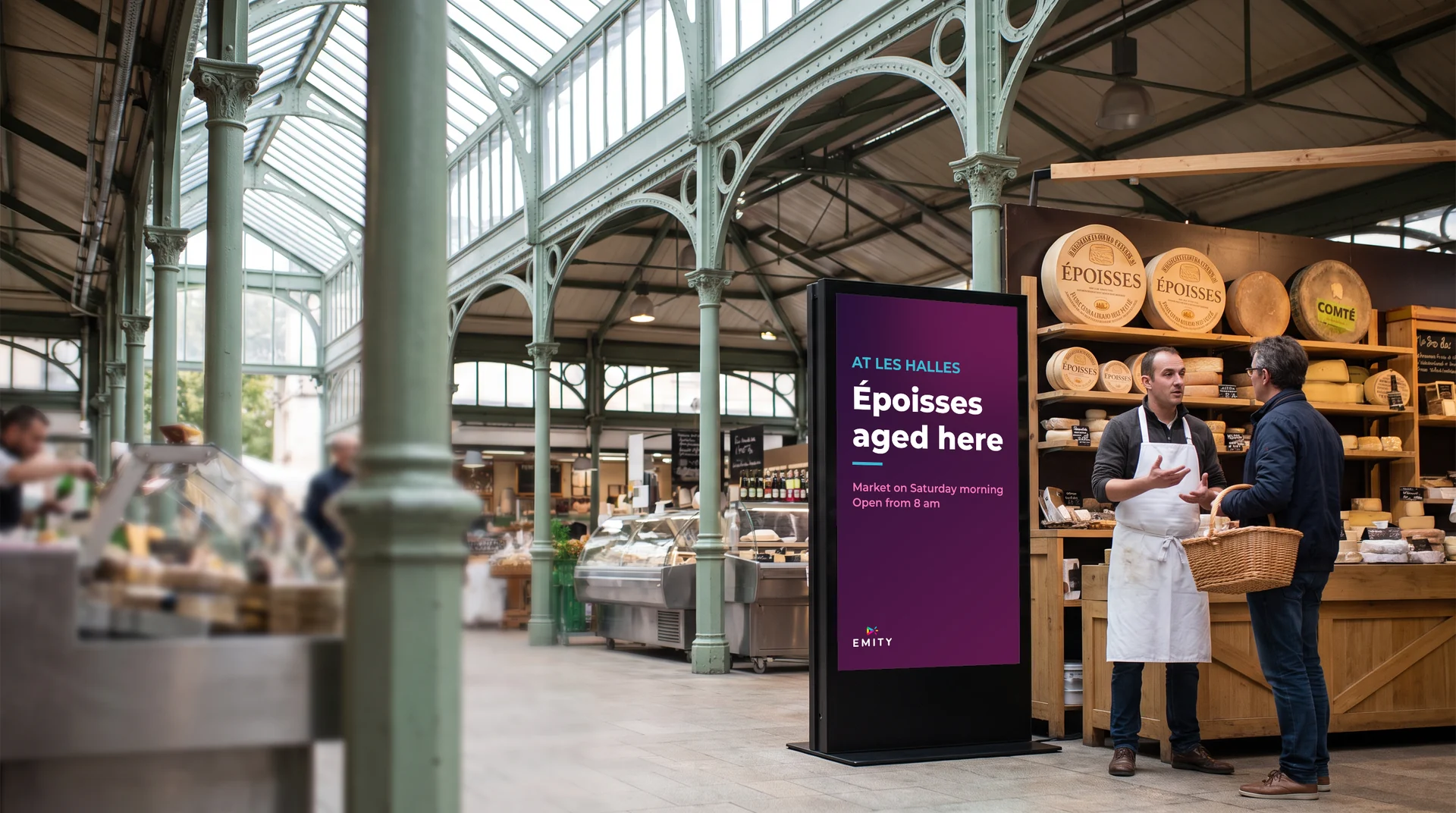 Les Halles in Dijon, cast-iron columns and glass roof: a cheesemonger advises a customer next to a totem reading “At Les Halles · Époisses aged here”