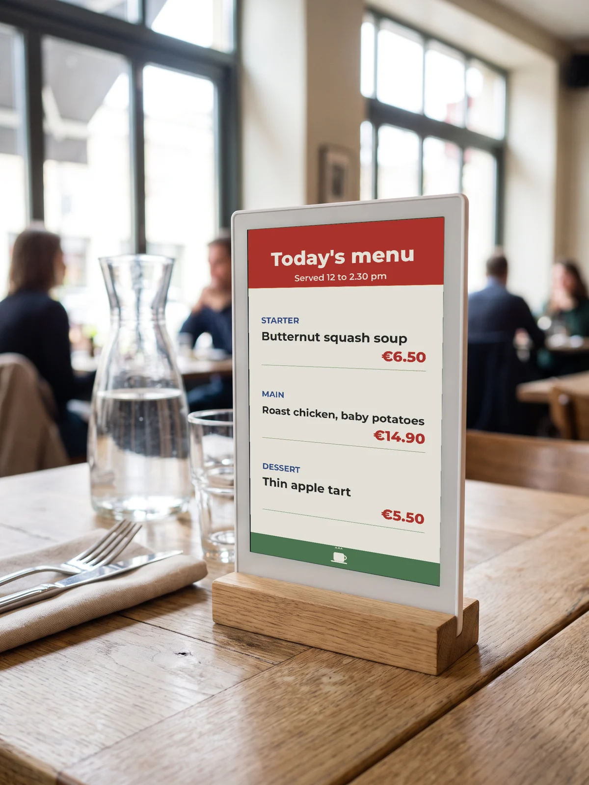 E-paper table stand showing “Today's menu” in a white frame on an oak base, on a bistro table next to a water carafe