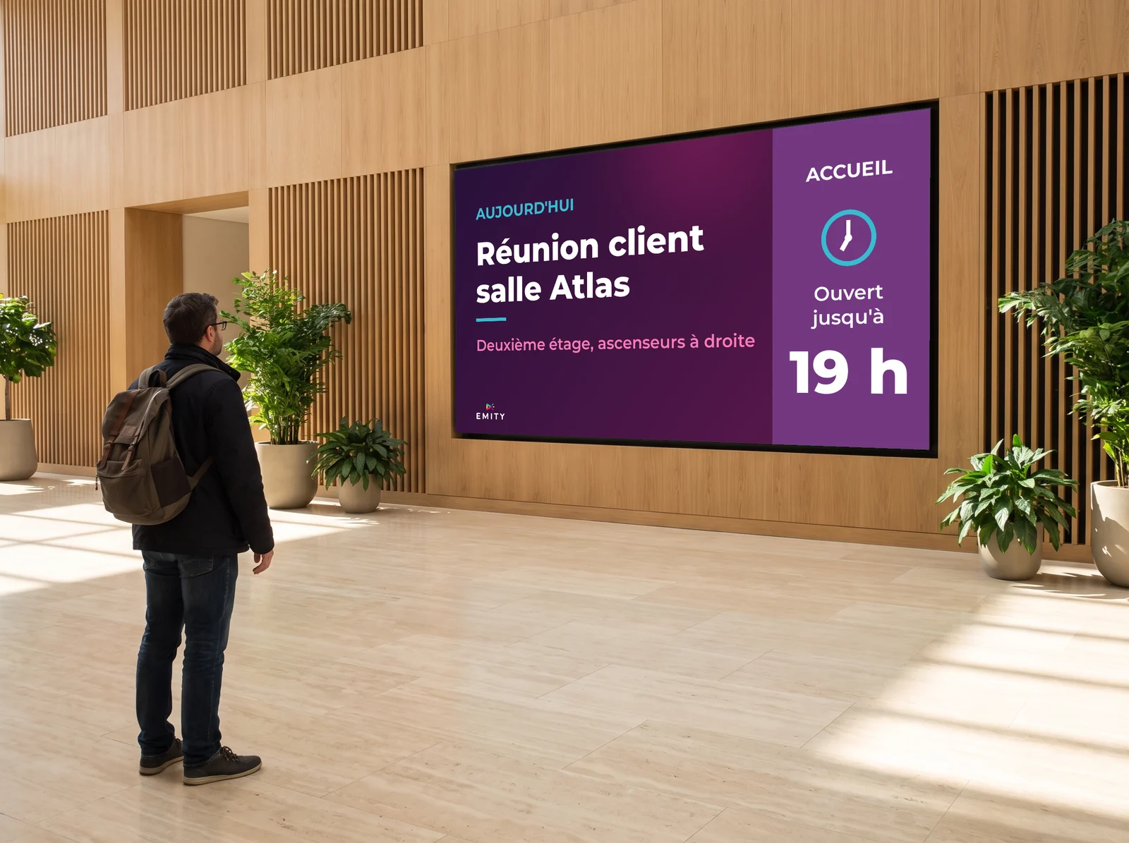 Visiteur arrêté à plusieurs mètres d'un grand écran mural dans un hall en bois clair : il lit la salle de sa réunion et l'étage sans s'approcher