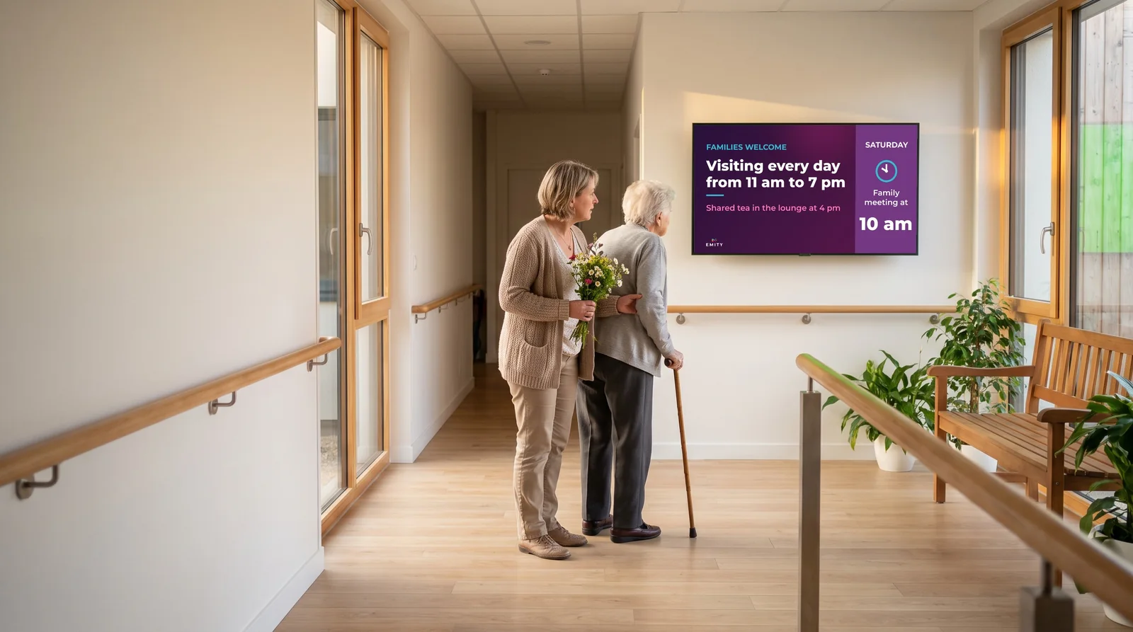 In a care home corridor, a daughter with a bouquet and her mother read the “Families welcome” screen: visiting hours and Saturday's family meeting