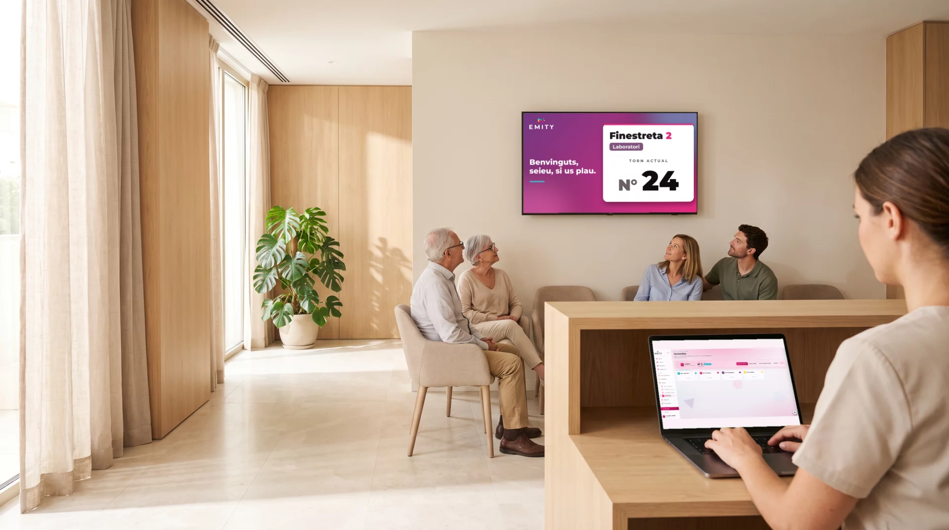 Sala d'espera d'un centre de salut: la pantalla mural crida el tiquet núm. 24 a la finestreta 2 mentre la recepcionista gestiona les crides a E-MANAGER