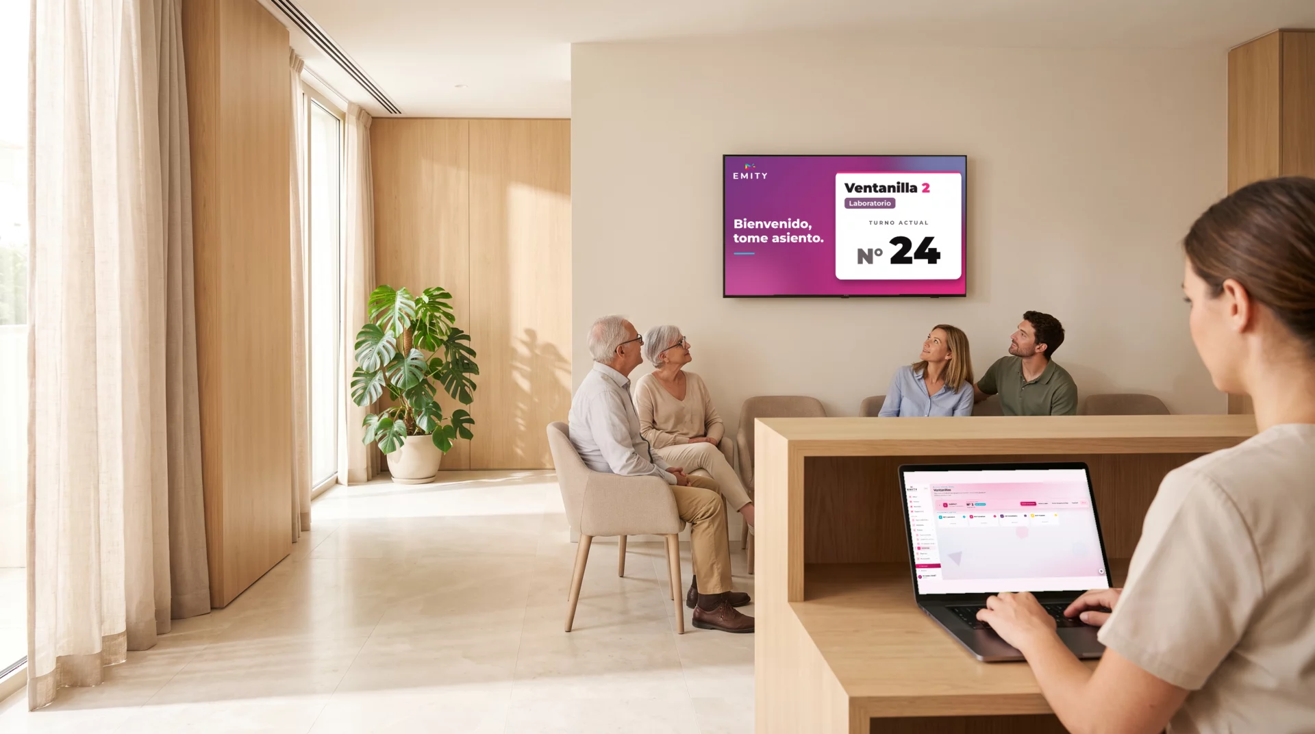Sala de espera de un centro de salud: la pantalla mural llama al ticket N.º 24 a la ventanilla 2 mientras la recepcionista gestiona las llamadas en E-MANAGER