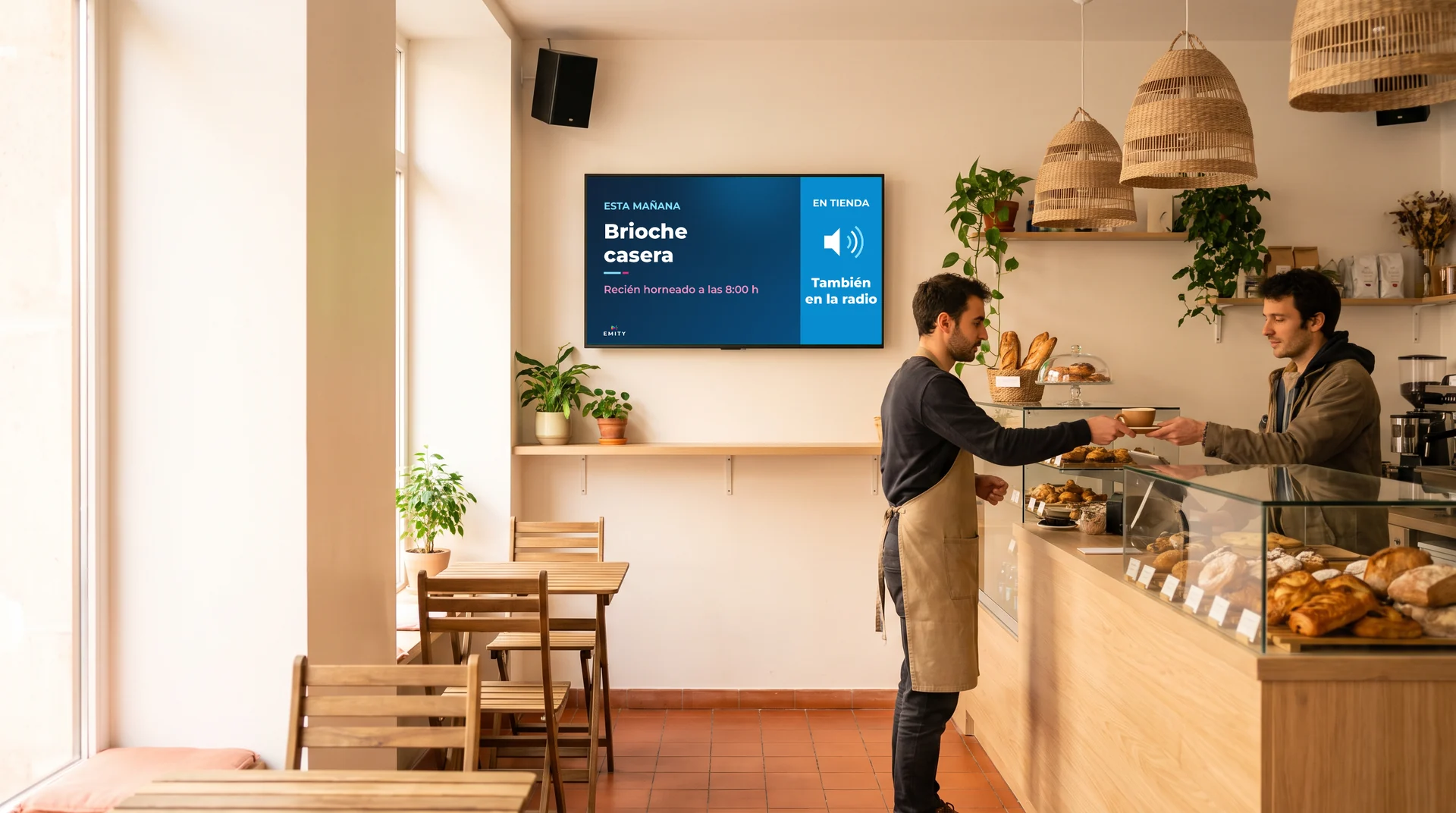 Panadería-cafetería de madera clara: un camarero sirve un café a un cliente bajo la pantalla « Esta mañana · Brioche casera · Recién horneado a las 8:00 h · También en la radio » y un altavoz de pared