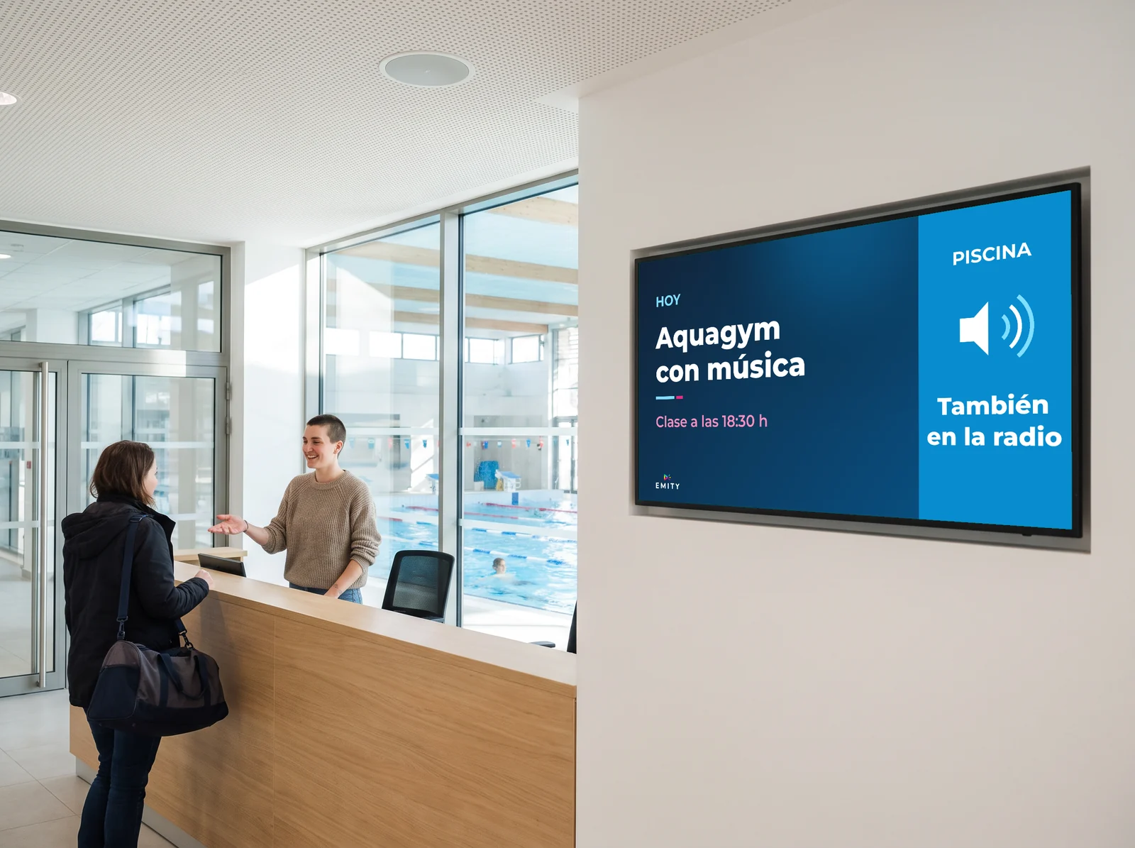Recepción de un centro acuático, piscina tras el ventanal: la pantalla « Hoy · Aquagym con música · Clase a las 18:30 h · También en la radio » junto al mostrador