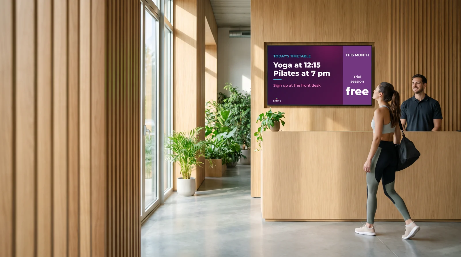 Club reception: a member looks up at the screen showing today's timetable, the coach behind the desk