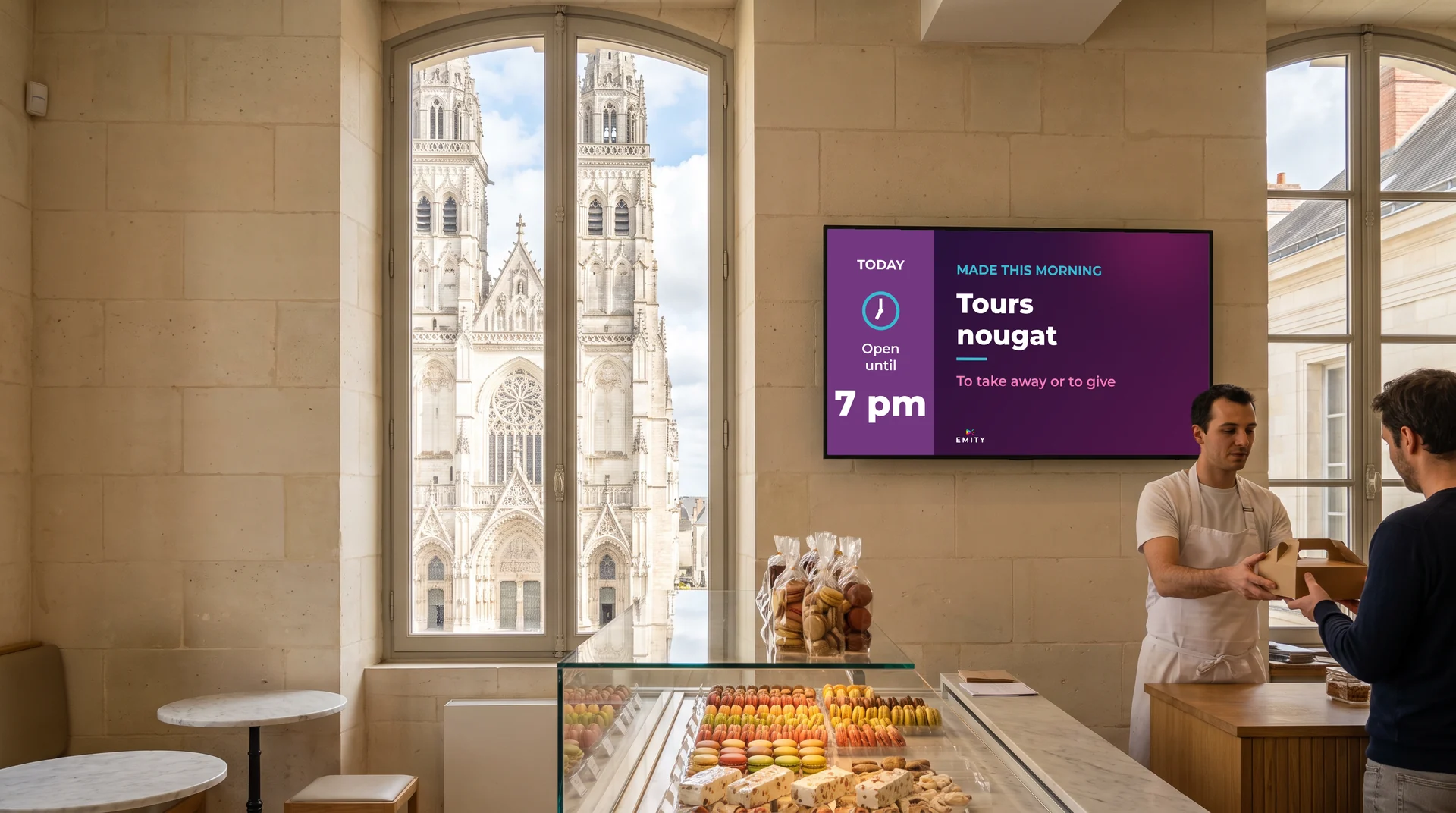 Pastry shop with tuffeau stone walls, the façade of Saint-Gatien cathedral through the window: a pastry chef hands a box to a customer beneath the wall screen “Tours nougat”